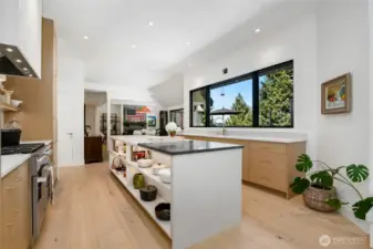 Designer kitchen with expansive center island, marble and soap stone countertops, open shelving, and sleek custom cabinetry. Oversized windows flood the space with natural light, offering picturesque views and a perfect blend of style and function.