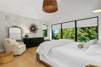 Serene main floor primary suite with vaulted ceilings, designer wallpaper, wide-plank hardwood floors, and expansive windows framing peaceful treetop and lake views.