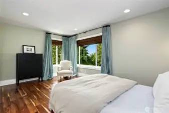 Sunlit second lower level bedroom retreat with rich hardwood floors, peaceful green-toned walls, and corner windows offering treetop views and tranquil natural light.