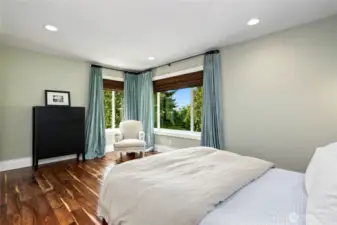 Sunlit second lower level bedroom retreat with rich hardwood floors, peaceful green-toned walls, and corner windows offering treetop views and tranquil natural light.