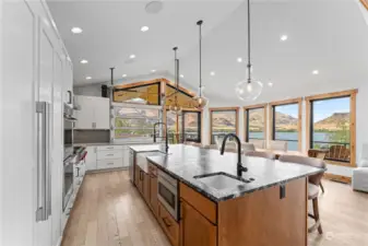 View of Main Level Kitchen w/ automated roll up garage door pass through, leather finish granite island w/ seating, high-end appliances, tile backsplash, dining room and glass sliding doors leading to the wraparound deck with outdoor kitchen, dining and lounge areas.