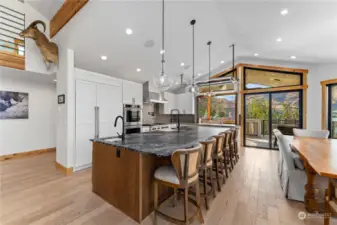 View of Main Level Kitchen w/ automated roll up garage door pass through, leather finish granite island w/ seating, high-end appliances, tile backsplash, dining room and glass sliding doors leading to the wraparound deck with outdoor kitchen, dining and lounge areas.