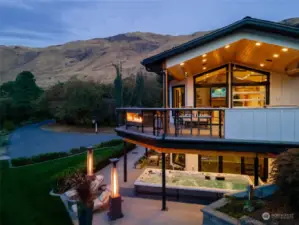 Twilight view of home w/ main level view deck w/ outdoor kitchen, dining area, see-through glass fireplace and lower level patio w/ hot tub/swim spa and seating area.