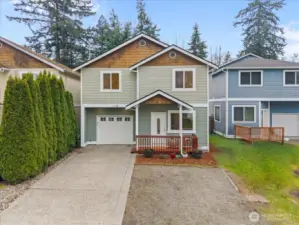 Front view showcasing the home’s covered entry porch with wood railing and landscaped planting area. Driveway and garage offer additional parking and storage.