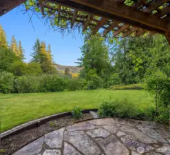 From beneath the vine-covered arbor, this view captures the expansive lawn and forested backdrop that define the home’s peaceful setting. The flagstone patio invites morning coffee or evening relaxation while overlooking mountain ridges in the distance. Framed by mature landscaping and surrounded by nature, the space feels like a private garden retreat—ideal for quiet moments, outdoor gatherings, or simply enjoying the beauty of the Pacific Northwest.