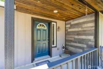 Covered front porch showcases a mix of rustic and modern finishes with a warm-toned tongue-and-groove ceiling, charcoal trim, and a feature wall with weathered wood-style paneling. The bold front door adds a pop of color, while the vertical sidelight window brings in natural light to the entry. Durable concrete flooring makes this entry practical for all seasons in the Pacific Northwest.