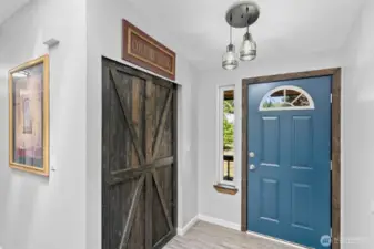 Bright and welcoming entryway featuring a statement barn-style sliding door, luxury vinyl plank flooring, and a bold blue front door with craftsman-style trim. A tall sidelight window adds natural light and frames the view of the front yard, while the modern farmhouse light fixture adds a touch of rustic charm. With neutral tones, custom millwork, and an efficient layout, this stylish foyer sets the tone for the rest of the thoughtfully updated interior.