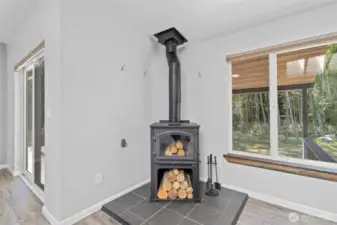 Efficient wood-burning stove offers both rustic charm and practical warmth, creating a cozy focal point for the main living area. Set on a tiled hearth and paired with built-in log storage and fireplace tools, this setup is ideal for chilly Pacific Northwest evenings. Large windows and nearby sliding glass doors frame peaceful wooded views, blending comfort and nature in one inviting corner of the home. Whether heating the space or adding ambiance, this feature enhances both sustainability and style.