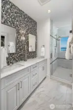 Elegant bath with dual vanity, marble countertops, designer tile accent wall, and glass-enclosed walk-in shower with island and water views.