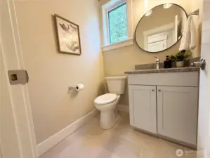 POWDER ROOM ON MAIN. This powder room presents a bright, clean, and thoughtfully designed space with a warm, welcoming feel. Soft neutral walls are paired with light tile flooring, creating an airy and cohesive look. A white vanity with simple, classic cabinetry is topped with a stone countertop, complemented by a round mirror and modern lighting that adds a subtle contemporary touch.