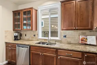 Large window above the stainless steel double sink fills the kitchen with natural light, complemented by full tile backsplash and elegant glass-front cabinetry.