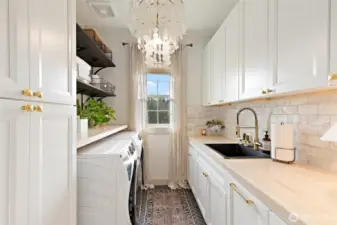 Laundry day just got a serious upgrade. This thoughtfully redesigned space features a 4.8 cu. ft. front-load washer with UltraFresh Vent System and Sanitize with Oxi, set beneath an extended butcher block counter for easy folding and organization. Soft-close white cabinetry, brushed gold hardware from Rejuvenation, and a marble tile backsplash elevate the entire room, while a built-in sink adds function to the form. Herringbone tile flooring and a timeless chandelier bring unexpected elegance, and open wood-stained shelving keeps essentials both accessible and stylish. A rustic sliding barn door completes the space, proving that utility can be just as beautiful as any other room in the home.