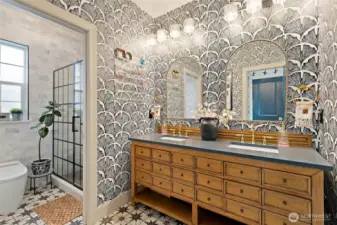 The main vanity area is a showstopper, featuring a double-sink layout with a warm, vintage-style wood cabinet and elegant gold fixtures. Bold, graphic wallpaper pairs beautifully with arched mirrors and statement lighting to create a dramatic yet inviting space. Patterned tile flooring ties everything together for a polished, high-design feel that elevates everyday routines.