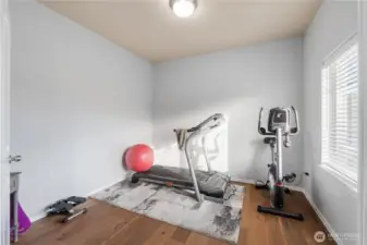 A bright flex room set up as a home gym, featuring a treadmill, stationary bike, and natural light from a large window. The soft wall color and warm hardwood floors create a calm, focused environment that can easily adapt to a home office, playroom, or guest space.