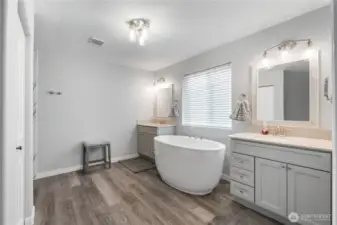 A spacious, updated primary bathroom featuring dual vanities, a sleek freestanding soaking tub, and a large window that fills the room with natural light. Soft gray cabinetry and modern lighting create a calm, spa-like atmosphere.