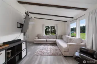 Comfortable living room with large windows offering peaceful views of the surrounding landscape. Features include vaulted ceilings with exposed beams, modern flooring, and plenty of natural light—ideal for relaxing or hosting guests.