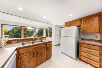Double-basin stainless steel sink is set beneath a wide bank of windows offering lush green views. This practical kitchen workspace combines functionality with a tranquil atmosphere.