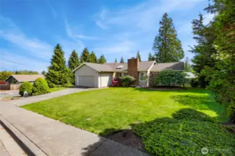 Sweeping corner-lot view of the home highlights a spacious front yard, privacy-enhancing evergreens, and a wide driveway. Thoughtful landscaping and natural surroundings offer both beauty and ease of maintenance.
