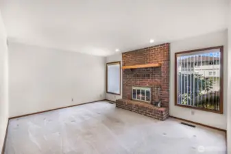 Spacious living room with a statement brick fireplace and wood mantel, flanked by large windows with vertical blinds that allow for privacy and light control. Clean carpet and recessed lighting complete the space.