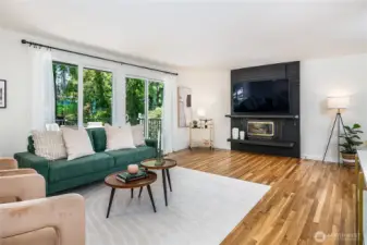 Bright and inviting main-level living room featuring a classic brick fireplace with gas insert, abundant windows, and a slider that opens to a spacious deck.