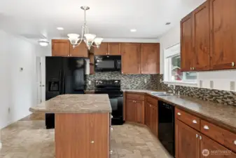 Here’s the kitchen showing off a little, with a glass tile backsplash that actually commits and runs the full height up to the cabinets on the range side and meets the windowsill on the sink side. A big vinyl window sits over the sink, shaker cabinets line the walls with brushed nickel pulls, and the appliances are coordinated in black. You get the French door fridge with water and ice, microwave and vent over the electric range, and the matching dishwasher. It’s a complete four-piece lineup, all staying, all ready for real life.