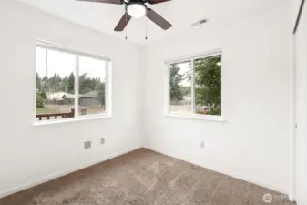 The front bedroom gets the corner treatment with twin windows and blinds, so there’s plenty of light without sacrificing privacy. White walls and latte-toned carpet keep it neutral, while the ceiling fan and ceiling-mounted heat register prove someone was actually thinking about comfort and furniture placement.