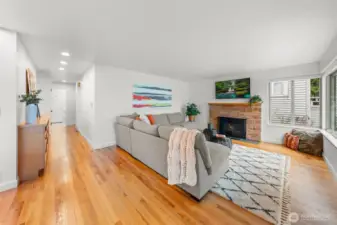 Expansive open living room anchored by a classic wood-burning fireplace with brick surround, creating a refined architectural focal point. Hardwood floors and generous natural light elevate the space for both everyday living and sophisticated entertaining.