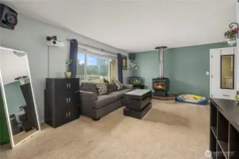 A bright and welcoming living room anchored by a charming wood-burning stove on a stone hearth. The expansive picture window not only floods the space with natural light but also provides a scenic view of the peaceful neighborhood beyond.