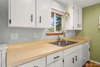 Close-up of the sink and prep area beneath the window. White upper and lower cabinets and simple laminate countertops offer easy-to-maintain surfaces with a timeless feel.