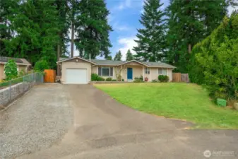 This wide-angle view highlights the home’s full frontage, offering a clear look at the extended driveway, attached single-car garage, and additional gravel parking space. The spacious front yard is framed by mature landscaping, with tall evergreens offering privacy and curb appeal.