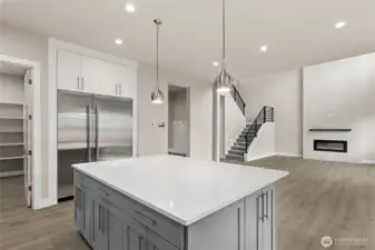 Spacious kitchen with quartz island and built-in refrigerator, offering direct views into the open great room with modern fireplace and sleek staircase. Walk-in pantry and mudroom access add everyday functionality to this elegant design.
