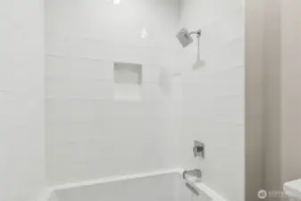 Close-up of the tiled shower and tub surround featuring oversized white subway tile, a built-in shampoo niche, and polished chrome fixtures. Clean, modern design with timeless appeal.