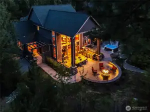 A stunning evening view of the outdoor kitchen, dining table, and firepit gathering area all softly illuminated for cozy evening entertaining. The home’s windows beam with warmth — a true reflection of comfort and lifestyle.