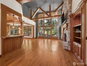 A dramatic open-concept great room with floor-to-ceiling windows, rustic wood beams, and a stunning stone fireplace. Custom built-ins flank the hearth, while natural light pours into the space overlooking outdoor living areas.