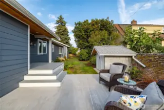 A view of the back yard from the patio, with brick fence running along the property line at right.  One of two storage sheds sits just off the patio.  Covered back deck leads into the mudroom.