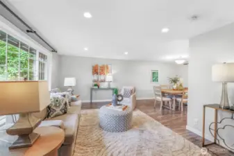 The spacious living room features a large picture window that fills the space with light and frames the greenery outside. The open floor plan creates a natural flow between living, dining, and kitchen areas.
