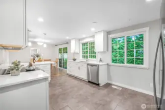 Large windows frame greenery outside, filling the kitchen with natural light. The layout provides both workspace and easy flow to the dining area and outdoor deck.