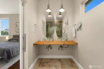 Modern elegance meets natural charm in this en suite bath, featuring dual glass vessel sinks, a custom live-edge vanity, and stylish oval mirrors under designer pendant lighting. The tile floor is an electric radiant heated floor.