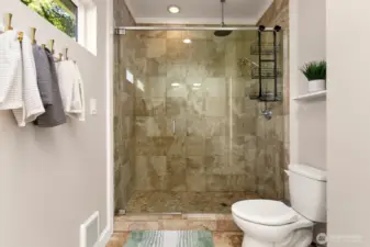 The primary suite features a spacious, walk-in tile shower with frameless glass doors, rainfall and handheld heads, and a pebble stone floor for a true spa-like experience.