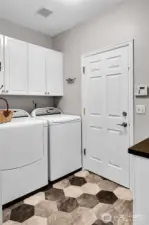 Light, bright laundry room. Paneled Quaker door. Whirlpool® Cabrio Platinum™ dryer and washer stay with home. Paneled white cabinets above. Folding counter with paneled white cabinet above and below. Faux-tile floor. Honeywell® security system. Step down to garage.