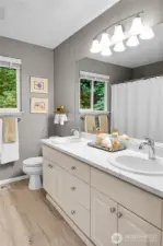 Main bath. Quartz counter with oval-shaped double sinks, chrome faucets. Mirror with bell shaped Alabaster style shades for vanity lights above. Window with screen two-inch blinds, looking out to the woods behind. Mansfield® standard watercloset. Bathtub with shower surround, privacy glass doors. Linen closet with tile floor, paneled Quaker door.