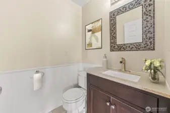 This stylish powder room on the main level features a modern vanity with a stone countertop and elegant mosaic-framed mirror. Crisp wainscoting and soft neutral tones create a clean, polished look that complements the home’s design.