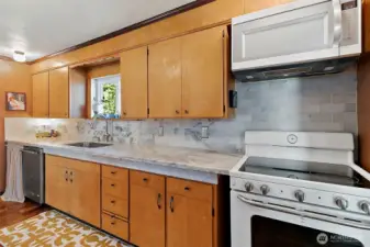 Galley kitchen with glass cook top, above range microwave, updated blacksplash and hardwood floors carrying through the house. Newer dishwasher sits to left of kitchen sink. Window above the sink and ample storage with top and bottom cabinetry.