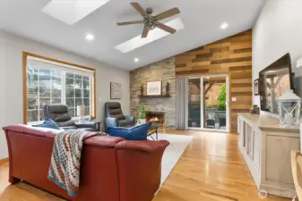 Spacious living room featuring a stone fireplace with a wood mantel, skylights, and recessed lighting. The room offers a comfortable blend of modern and rustic elements, with hardwood floors, large windows, and access to the backyard through sliding doors. A perfect space for relaxing or entertaining.