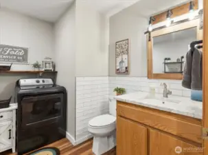Combined bathroom and laundry area with vanity, quartzite countertop, and front-loading dryer. Open shelving provides additional storage.