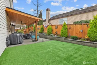 Patio area with seating space, potted plants, and raised garden beds along the fence line. Artificial turf and stone accents create a low-maintenance yard.