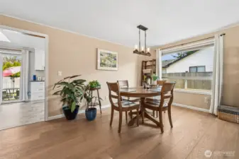 The dining room shows up ready to host everything from casual takeout nights to full holiday spreads. It’s a generously sized space anchored by those beautiful medium tone engineered wood floors that bring warmth and a timeless feel underfoot. A huge picture window pulls in natural light and gives the room an airy, open presence that makes it easy to dress up or keep relaxed. Overhead, the contemporary chandelier makes a confident style statement with its sleek wood bar design and clear glass lights suspended by clean chain details. From this vantage point you can see right into the kitchen and even catch a glimpse of the slider leading out to the backyard, which keeps the whole main living area feeling connected and social in the best way.