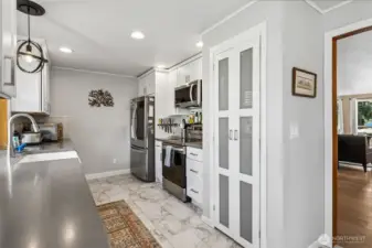 This galley kitchen is a quiet overachiever. Everything is right where you want it, creating that smooth, step saver flow that makes cooking feel efficient and surprisingly enjoyable. Newer flooring keeps things fresh underfoot, while a long stretch of hard surface counters gives you plenty of room to prep, spread out, and maybe even stage your next signature dish. Crisp white shaker cabinetry keeps the look timeless, paired with the softest dove gray walls that add just enough warmth. Stainless appliances include a sleek French door fridge with bottom freezer and door dispenser, plus a clever frosted glass pantry closet that keeps snacks, staples, and small appliances easily within reach while still looking polished.