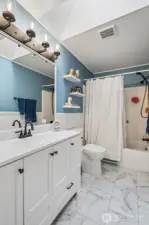 The main bathroom leans classic with a little personality twist. Beadboard wainscoting grounds the space while the soft stormy blue walls bring just enough mood to keep it interesting. A long vanity gives everyone elbow room for those busy morning routines, and the full tub setup features a dual showerhead with handheld sprayer for extra flexibility. The sleek skirted style toilet makes cleaning feel less like a chore, and overall the room lands in that sweet spot of practical and polished.