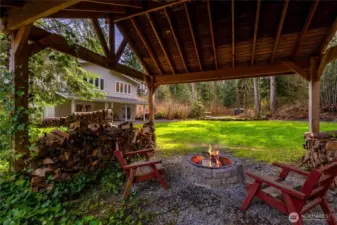 Step outside to a covered timber-frame pavilion, where a built-in fire pit creates the perfect gathering space year-round. Surrounded by stacked firewood and lush greenery, this area is ideal for entertaining or unwinding under the canopy of trees.